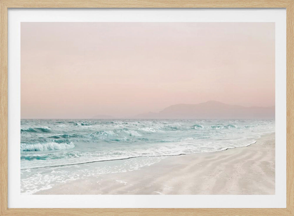 A serene coastal scene with gentle turquoise waves washing onto a sandy beach. The sky is a soft, hazy pink, and faint outlines of mountains are visible in the distant background. The artwork is presented in a silver frame. Print