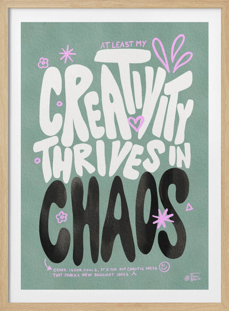 A motivational art print with a sage green background featuring the quote 'AT LEAST MY CREATIVITY THRIVES IN CHAOS'. The words 'CREATIVITY' and 'THRIVES IN' are in a large, bubbly white font, while 'CHAOS' is in a bold black font. The design is accented with small pink doodles like flowers, stars, and hearts. A smaller quote at the bottom reads, 'ORDER IS FOR FOOLS, IT'S THE HOT CHAOTIC MESS THAT SPARKS NEW BRILLIANT IDEAS'. Wall Art