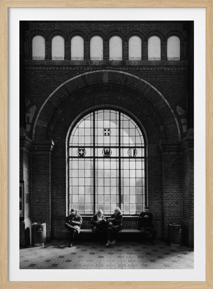 A dramatic black and white photograph of a large, brick-walled waiting area, possibly a train station. Four people are seated on a long bench beneath a massive arched window, which dominates the scene. The lighting creates a moody, contemplative atmosphere, highlighting the grandeur of the architecture. Artwork