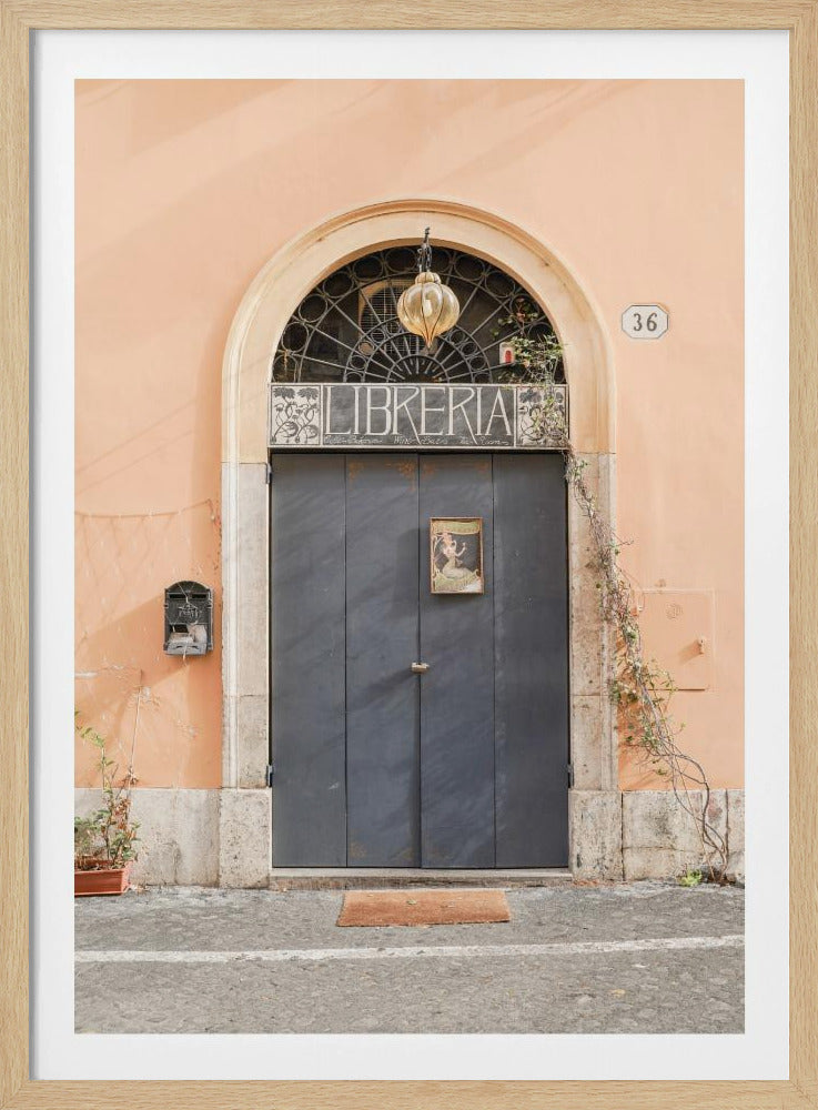 A charming European bookstore entrance featuring a large, dark grey arched door set against a peach-colored wall. Above the door, a sign reads 'LIBRERIA' below a decorative fanlight window and a hanging lantern. Decor