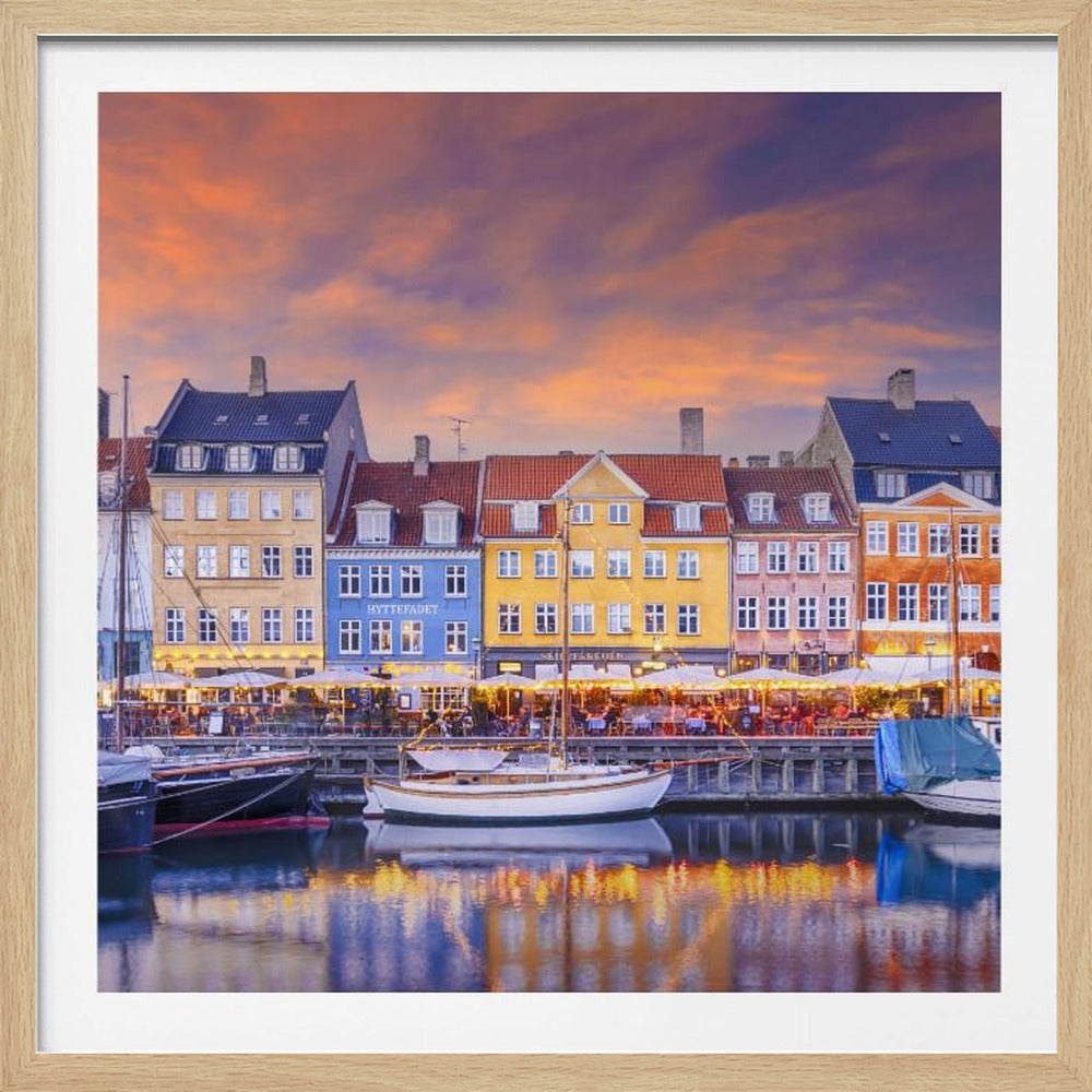 A framed photograph of the Nyhavn harbor in Copenhagen at dusk, featuring a row of brightly colored historic buildings along the canal. Sailboats are moored in the water, which reflects the warm glow of the building lights and the vibrant orange and purple sunset sky. Print