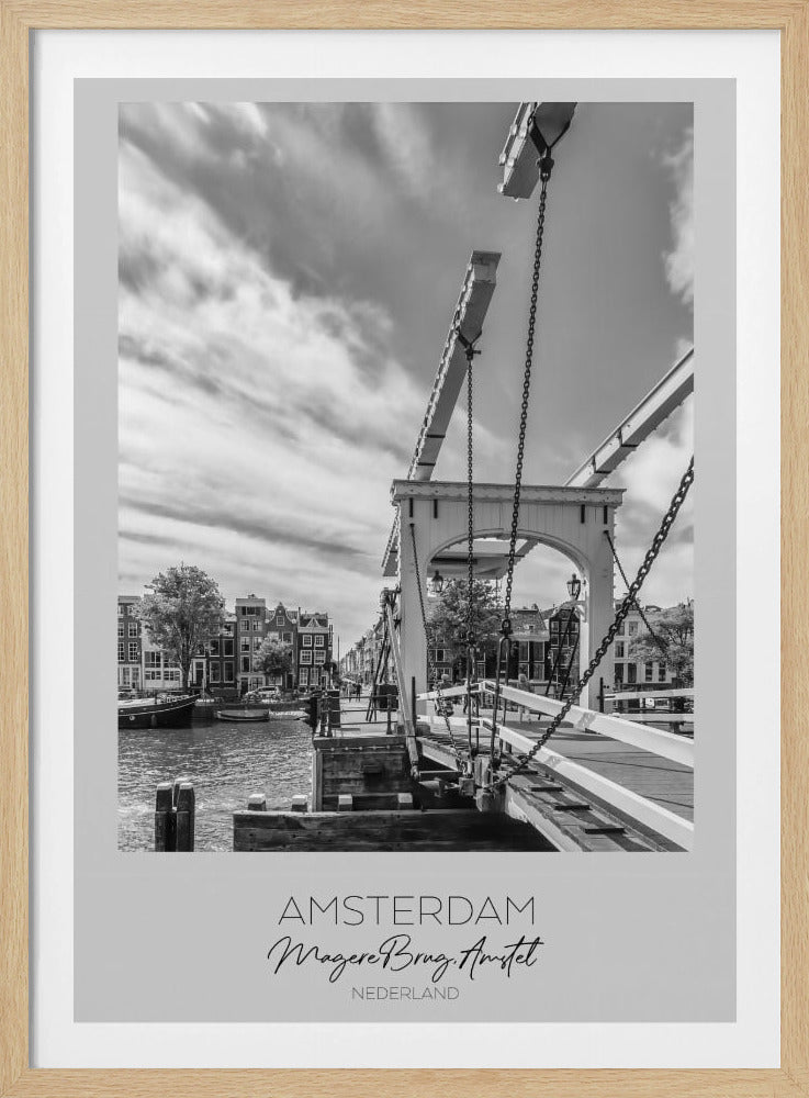 A black and white travel poster of Amsterdam, featuring a side view of the Magere Brug, a traditional wooden drawbridge, over the Amstel river. Classic canal houses line the waterway in the background under a dramatic, cloudy sky. The image is in a black frame and has text at the bottom that reads 'AMSTERDAM', 'Magere Brug, Amstel', and 'NEDERLAND'. Artwork