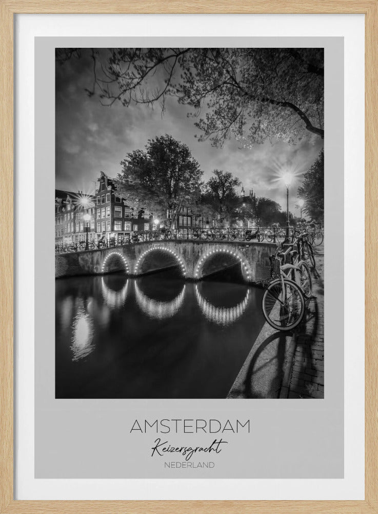 A black and white poster of Amsterdam's Keizersgracht canal at night. An arched bridge with glowing lights is the centerpiece, its reflection shimmering on the dark water. Bicycles are parked along the bridge and in the foreground on a cobblestone path. Classic canal houses and trees line the background under a dramatic, cloudy sky. The image is framed with text below reading 'AMSTERDAM Keizersgracht NEDERLAND'. Artwork