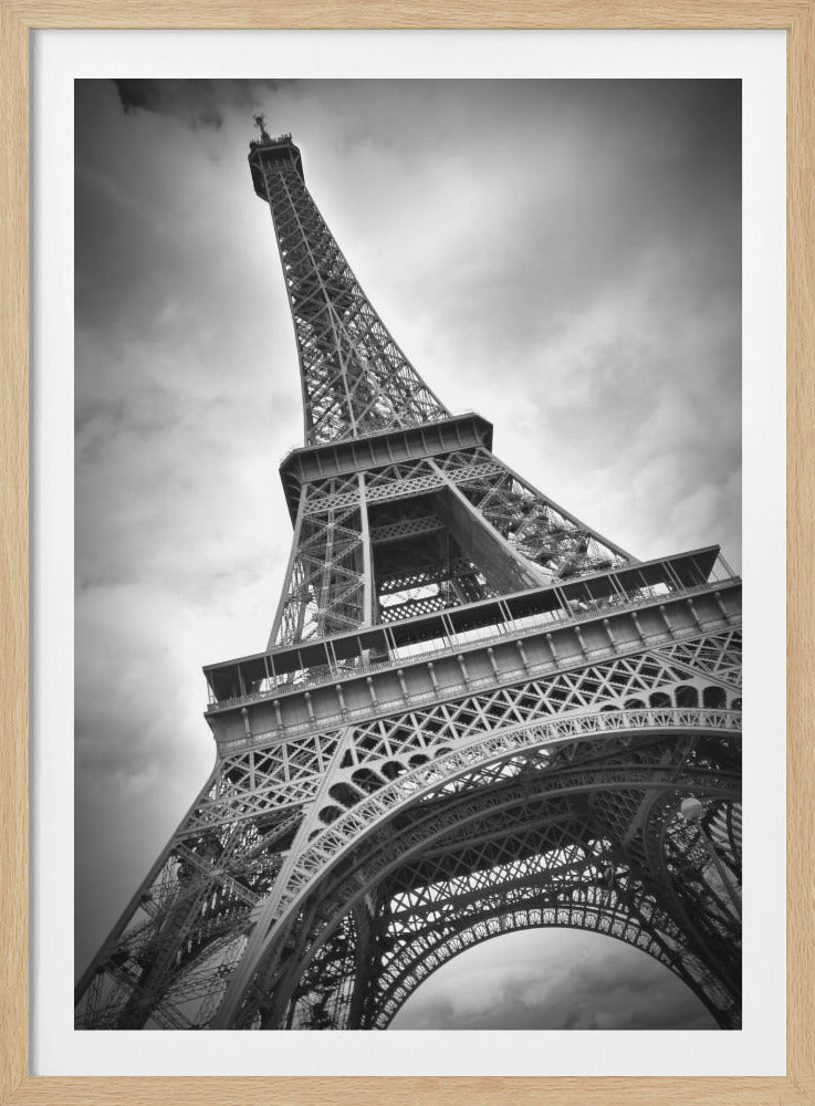 A dramatic, low-angle, black and white photograph of the Eiffel Tower, looking up from near its base. The intricate iron lattice structure fills the frame against a backdrop of a cloudy, moody sky, emphasizing its grand scale and architectural detail. Artwork