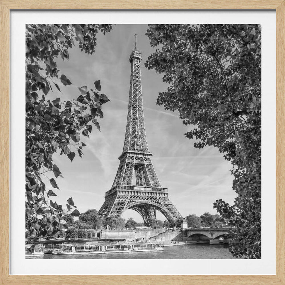 A framed, black and white photograph of the Eiffel Tower in Paris. The view is from across the Seine River, with the tower framed by the leaves and branches of trees in the foreground. Tour boats are visible on the water near the riverbank. Wall Art