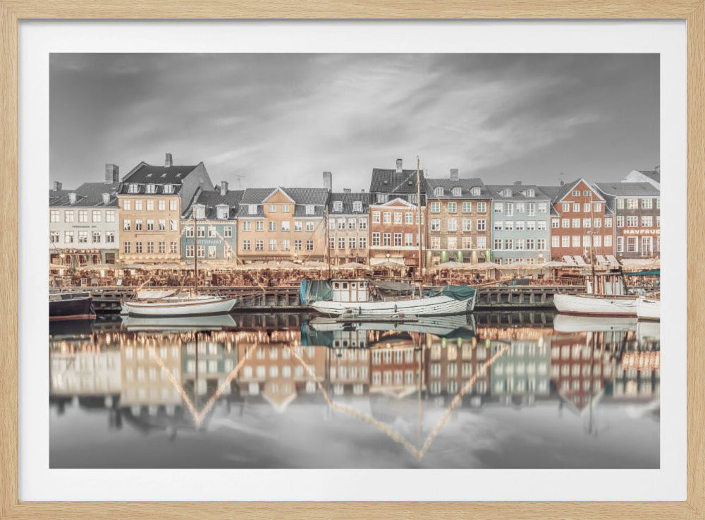 A framed, color-selective photograph of the Nyhavn harbor in Copenhagen. A row of colorful 17th and 18th-century townhouses lines the waterfront, with their warm lights and facades reflecting in the calm canal. The sky and parts of the water are in black and white, making the colored buildings stand out. Several boats are moored along the quay. Decor