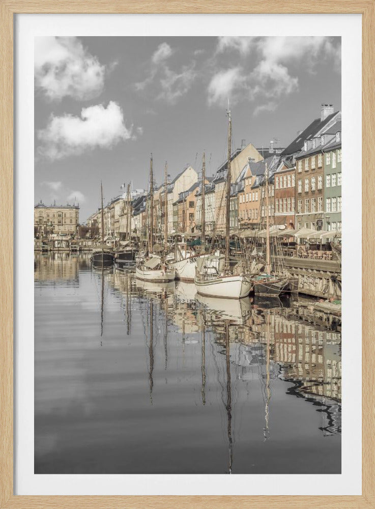 A muted, desaturated photograph of the Nyhavn harbor in Copenhagen. Old-fashioned sailboats are moored along the canal in front of a row of colorful historic buildings. The boats, buildings, and a cloudy sky are perfectly reflected in the calm water. Wall Art