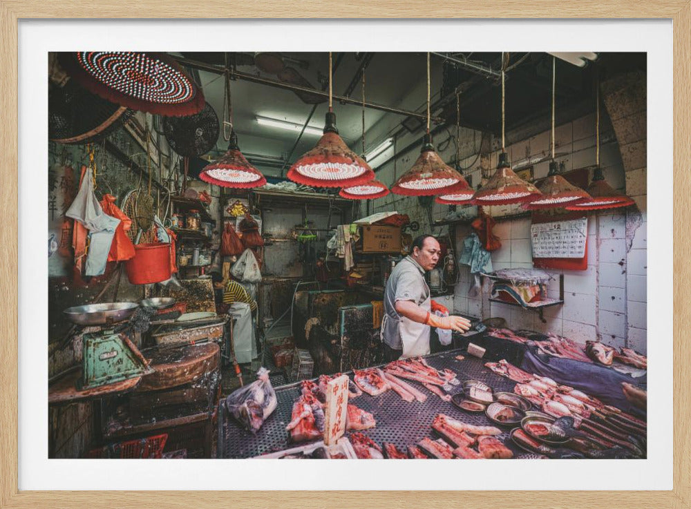 A candid photograph of a butcher working at an indoor Asian street market, with various cuts of raw meat and fish displayed on a metal counter under the warm glow of several large, red-rimmed hanging lamps. Artwork