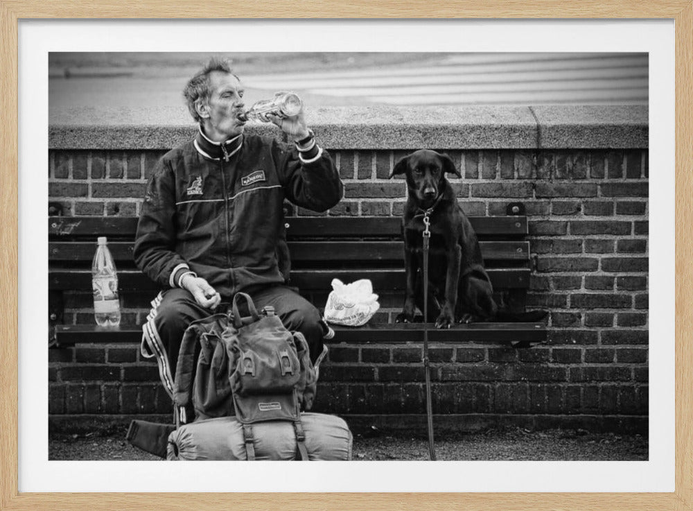 A framed, black and white photograph of a weary man and his loyal black dog sitting together on a park bench. The man, with a large backpack and bedroll at his feet, is drinking from a water bottle, while the dog sits upright, looking forward. Wall Art