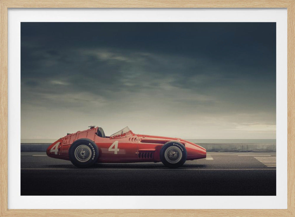 A side profile of a vintage red single-seater race car with the number 4 painted on it, parked on a seaside road under a dramatic, cloudy sky. The image has a muted, nostalgic color palette and is shown within a silver frame. Decor