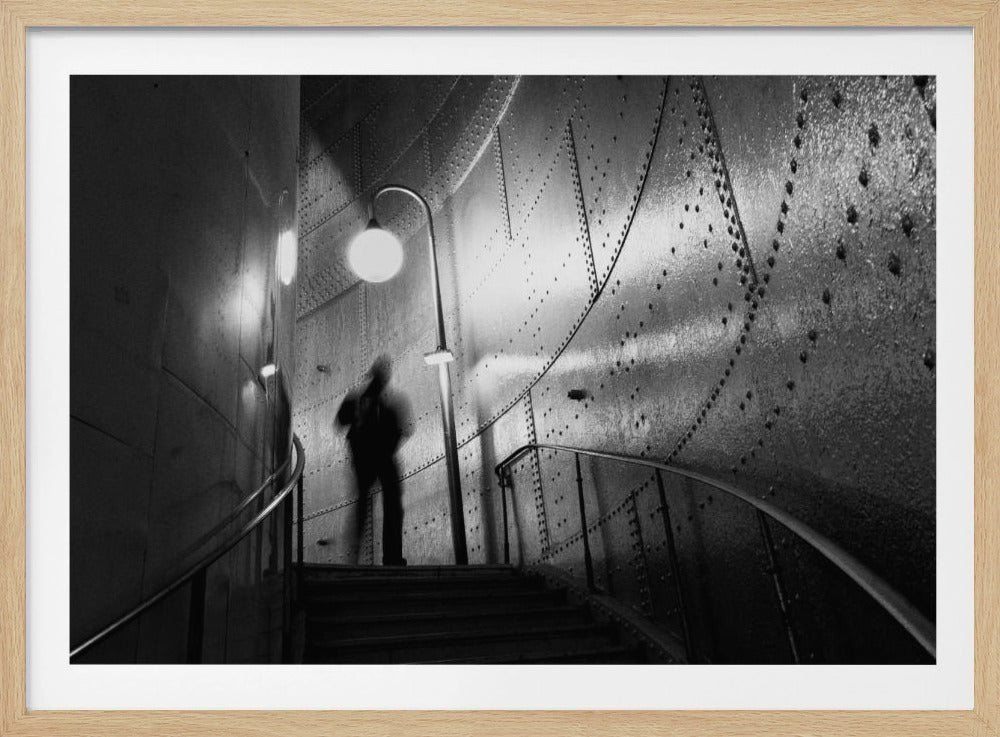 A moody, black and white photograph showing a blurred silhouette of a person walking up a staircase. The scene is illuminated by a single street-style lamp, casting light and shadows on the curved, riveted metal walls of the industrial-looking interior. Decor