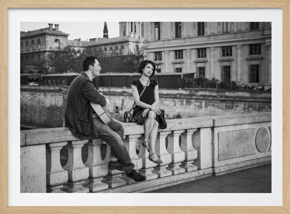 A romantic black and white photograph, set in a silver frame, showing a man playing an acoustic guitar for a woman as they sit together on a stone bridge balustrade in a European city. Poster