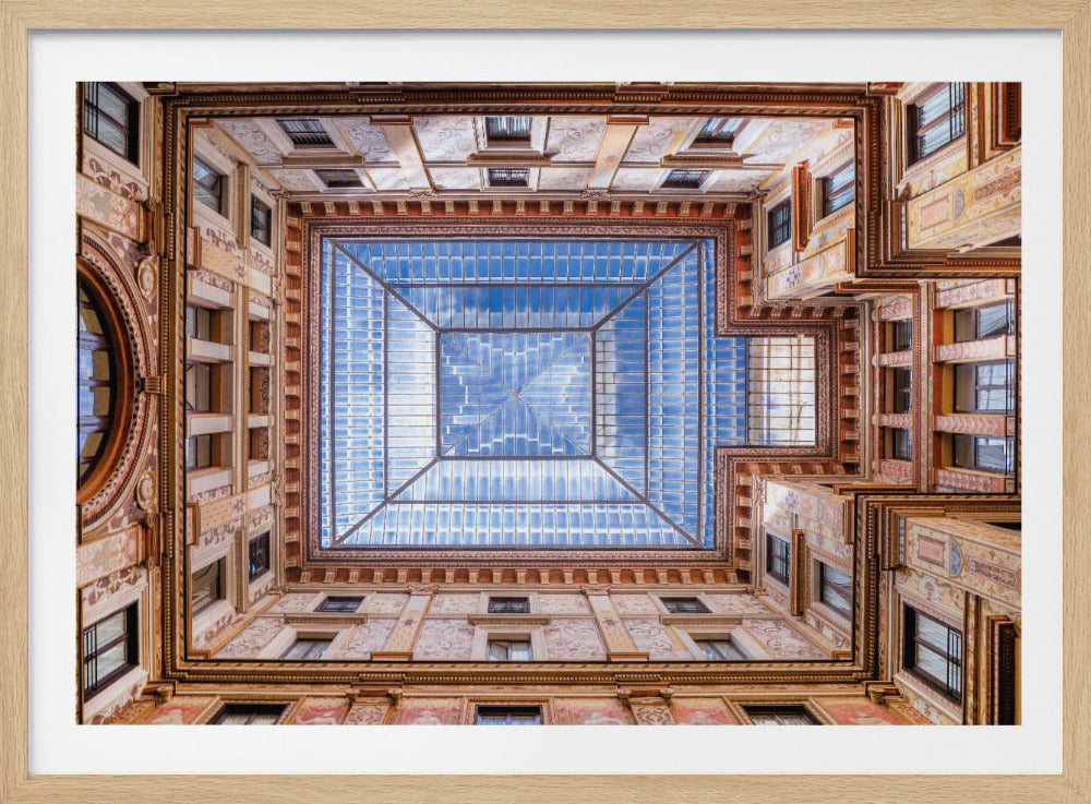 A worm's-eye view looking straight up from the center of an ornate, multi-story courtyard. The detailed walls create a symmetrical frame around a large glass skylight, which reflects a bright blue sky with fluffy white clouds. Print