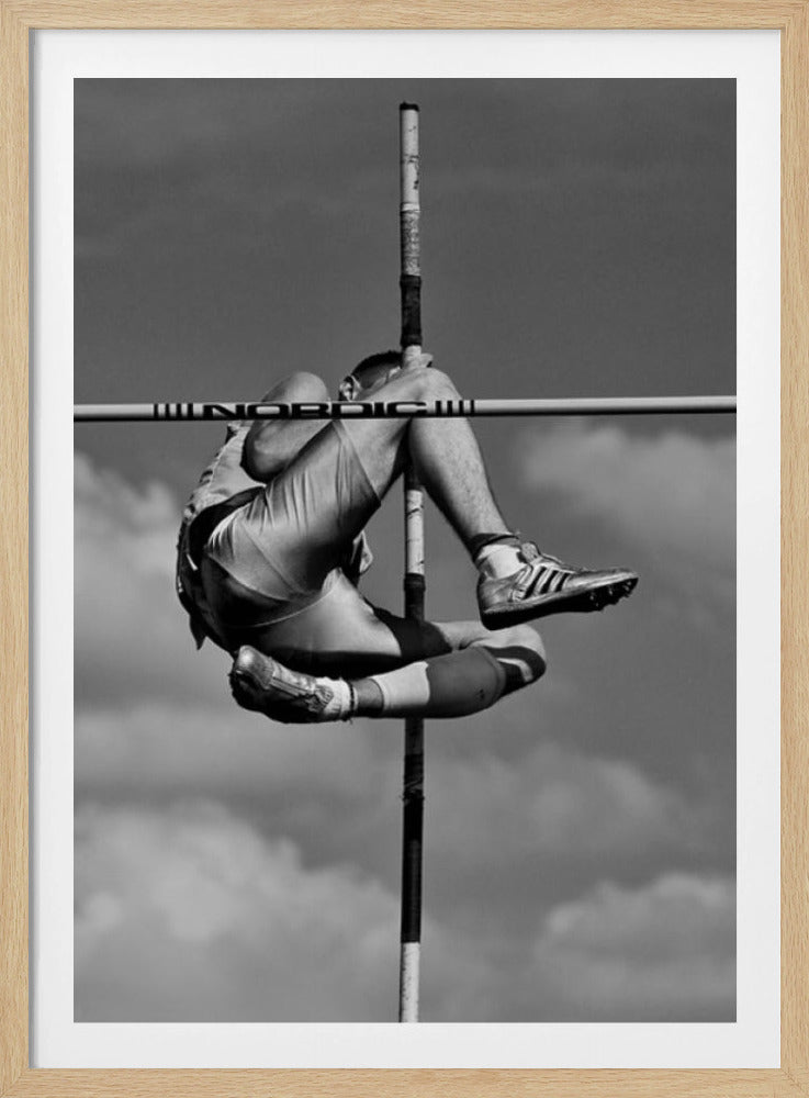 A dramatic black and white, low-angle photograph of a male athlete in mid-air during a pole vault. He is arching his back and bending his legs to clear the horizontal bar, with the vertical pole bisecting the frame. The background is a partly cloudy sky. Wall Art