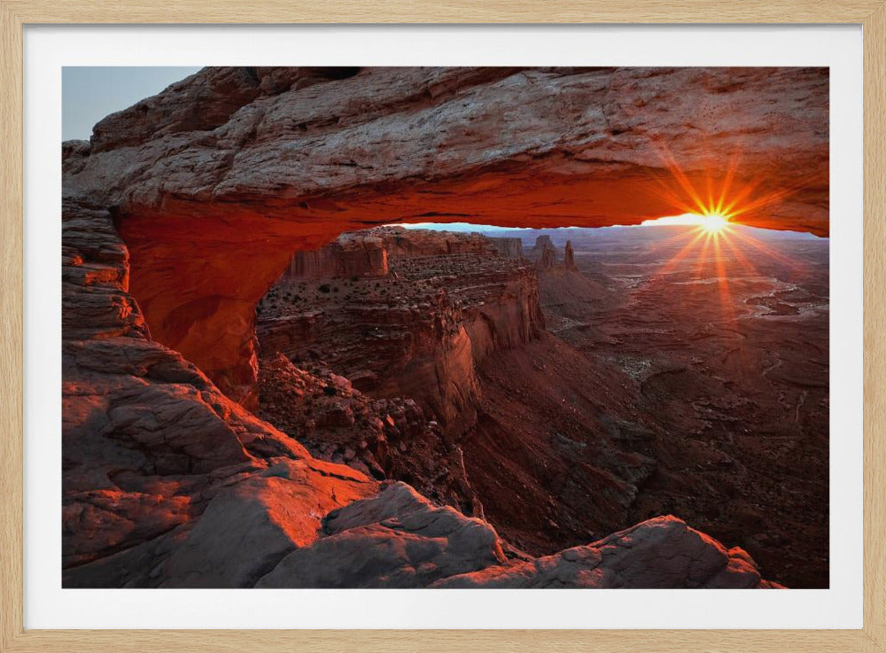 A spectacular sunrise seen through the natural frame of Mesa Arch in Canyonlands National Park, Utah. The sun bursts over the horizon, casting a dramatic orange and red glow on the underside of the rock arch and the vast canyon landscape below. Poster