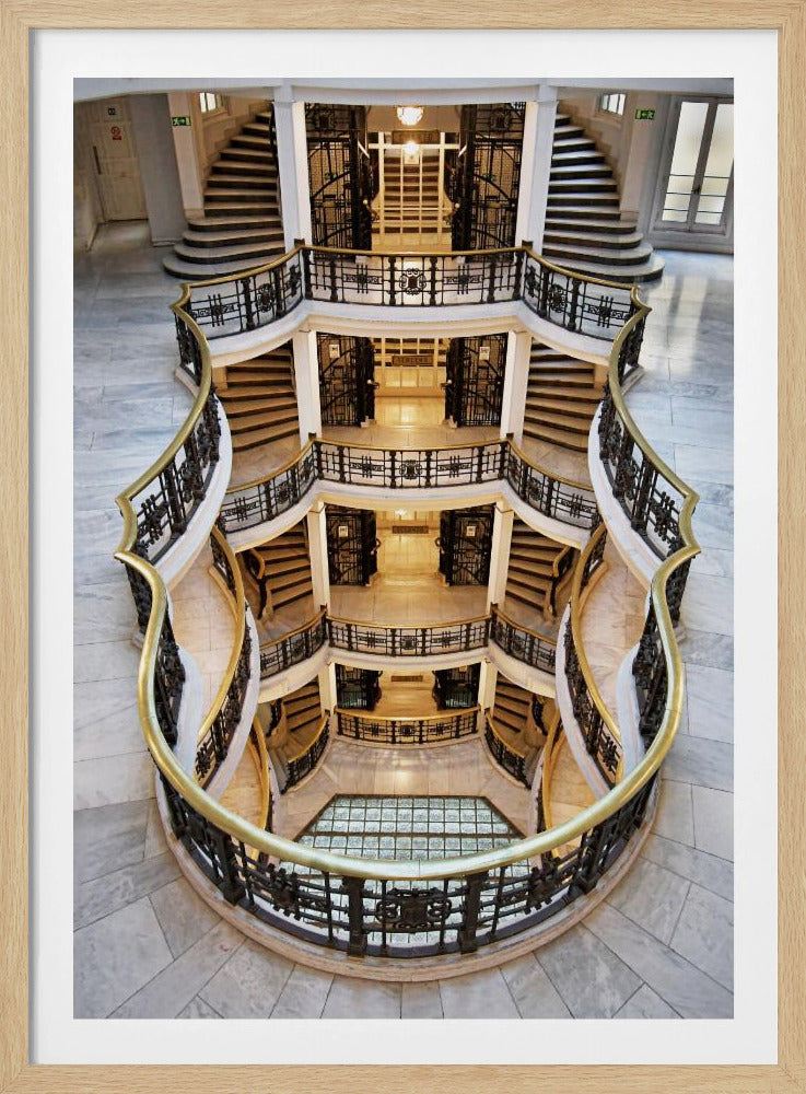 A high-angle photograph looking down into the atrium of a luxurious building, showcasing a symmetrical, multi-level, curving staircase with ornate black ironwork and gleaming gold handrails on white marble floors. Poster