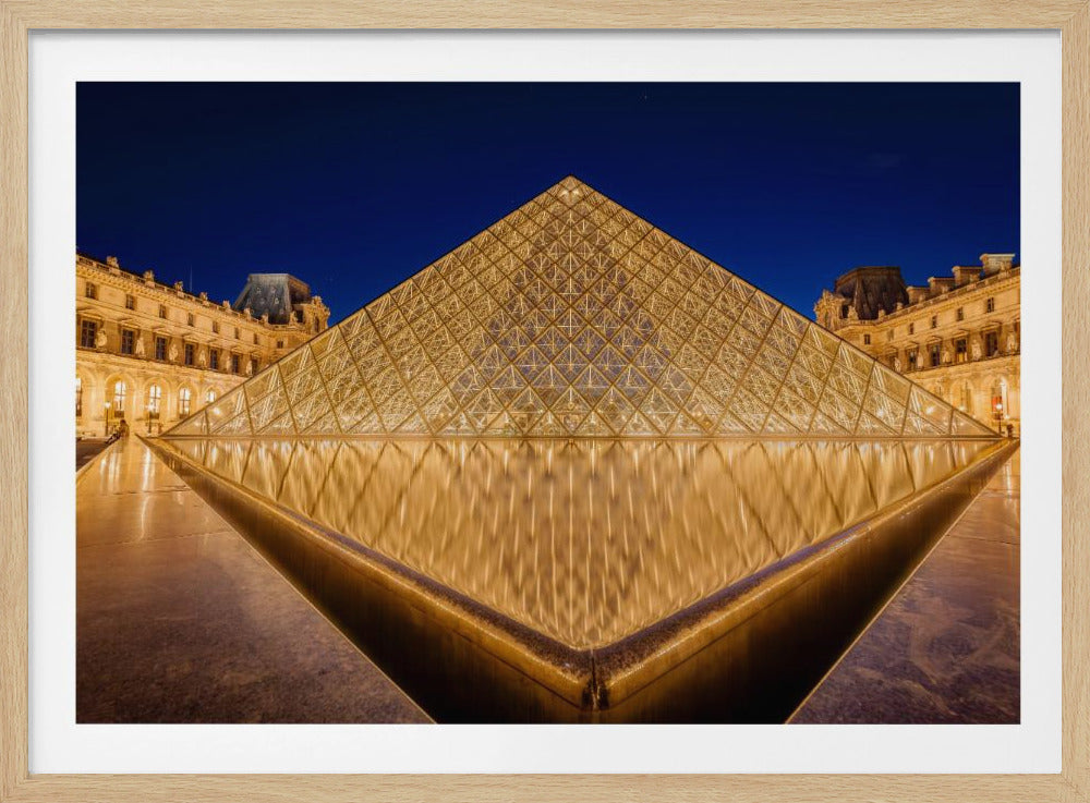 A low-angle, symmetrical night photograph of the illuminated glass Louvre Pyramid in Paris. The pyramid glows with a warm golden light against a deep blue night sky, and its perfect reflection is visible in the still water of the fountain in the foreground. The historic Louvre Palace is visible in the background. Print