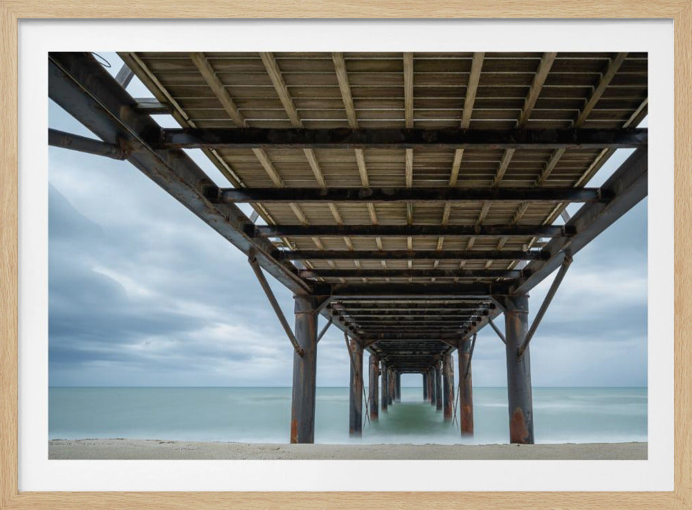 A symmetrical, low-angle photograph taken from underneath a long pier, looking out to sea. The rusty metal pilings and wooden planks of the pier create a receding perspective over the smooth, long-exposure water, all under a cloudy sky and presented within a silver frame. Poster