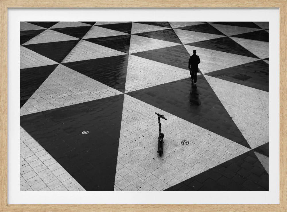 A high-angle, black and white photograph of a man in a coat and fedora walking away across a vast plaza with a geometric pattern of black and light gray triangles. An electric scooter stands alone in the foreground, creating a sense of solitude and contrast. The image is presented within a silver frame. Decor