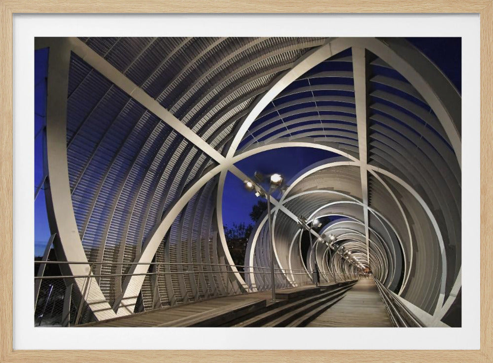 A long perspective view from within a modern, metallic pedestrian bridge at night. The structure is composed of swirling, intersecting silver arches that create a tunnel-like effect, illuminated by streetlights against a deep blue evening sky. The image is presented within a silver picture frame. Artwork