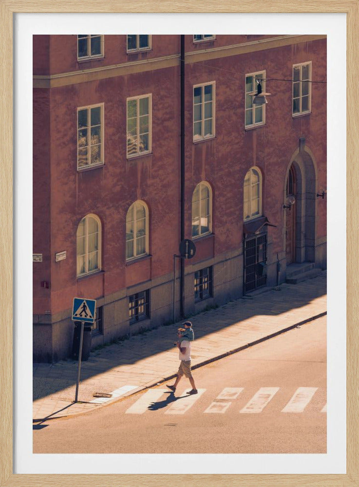 A high-angle photograph of a man carrying a child on his shoulders as they walk across a sunlit crosswalk, with a large, reddish-brown European-style building in the background. The scene is bathed in warm light, creating long shadows. Poster