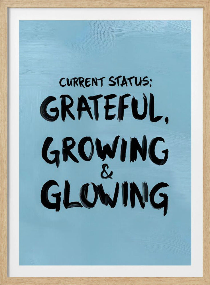 A motivational poster with a light blue background and a thin black frame. The text 'CURRENT STATUS: GRATEFUL, GROWING & GLOWING' is written in a bold, black, handwritten brush font. Decor