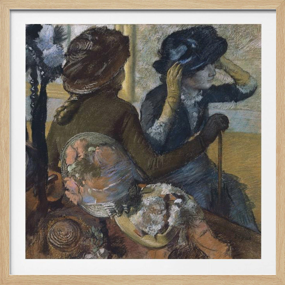 A framed impressionist painting by Edgar Degas, 'The Millinery Shop', depicting two women. One woman, seen from the back, wears a brown coat and hat, while the other tries on a dark blue hat, adjusting it with her gloved hands. Colorful hats are displayed in the foreground. Print