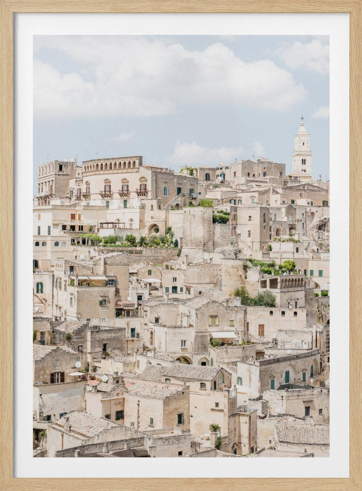 A vertical photograph of the ancient, stone city of Matera, Italy, with its dense collection of beige buildings and tiled roofs built up a hillside under a pale blue sky. A white bell tower stands out against the skyline. Poster