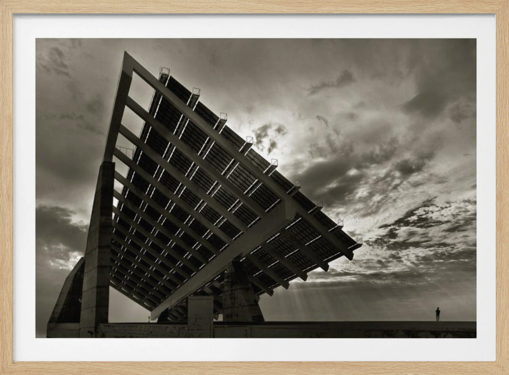 A framed, dramatic black and white photograph showing a massive, tilted solar panel structure from a low angle. The sky is filled with dark, moody clouds with some sun rays breaking through. A tiny silhouette of a person in the lower right corner emphasizes the grand scale of the architecture. Wall Art