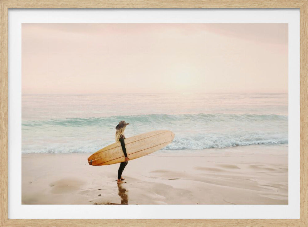 A female surfer with long blonde hair, wearing a black wetsuit and a hat, holds a surfboard while standing on a wet sandy beach looking out at the ocean during a hazy pink sunset. Decor
