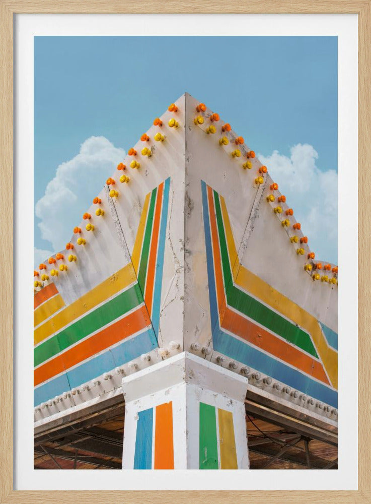 A low-angle photograph of the corner of a retro-style building against a bright blue sky with white clouds. The white facade is painted with bold, colorful stripes in orange, yellow, green, and blue, and the angled roofline is decorated with rows of unlit yellow and orange carnival lights. Print