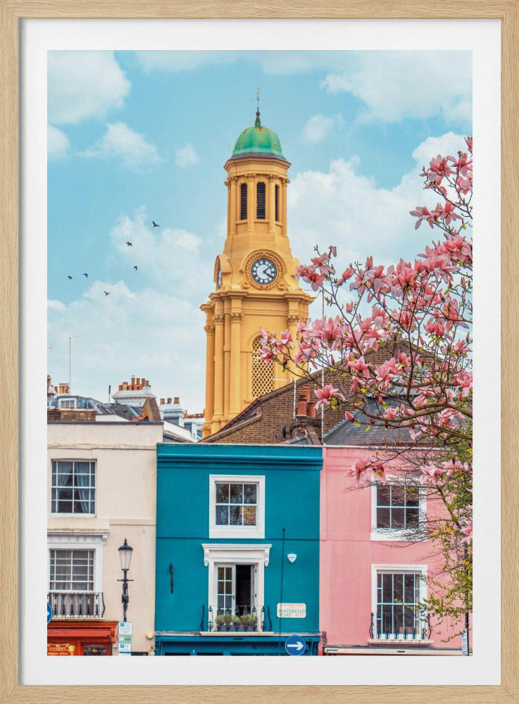 A vibrant city street scene on a sunny day, featuring a row of colorful houses in teal, pink, and off-white. A striking yellow clock tower with a green dome stands tall against a bright blue sky, with a blooming pink magnolia tree in the foreground. Print