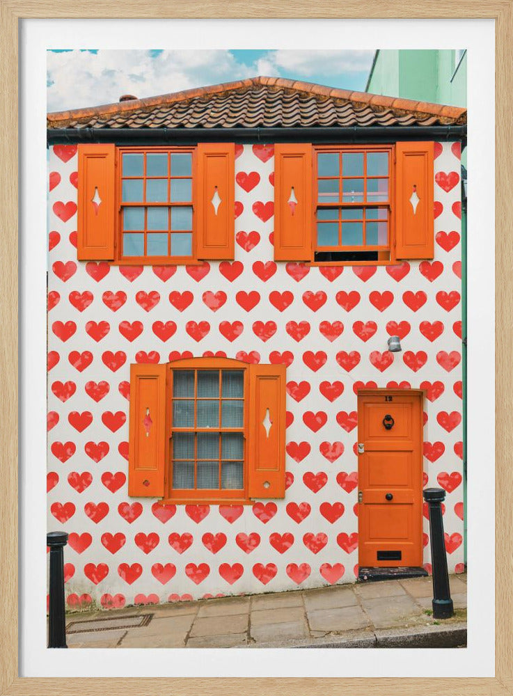 The front facade of a whimsical two-story house with white walls covered in a pattern of red hearts. The house has bright orange shutters on its windows and a matching orange front door, creating a vibrant and playful look against the heart-patterned background. Artwork
