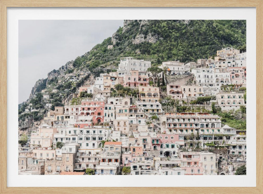 Photograph of the iconic cliffside village of Positano on the Amalfi Coast, showcasing a dense cascade of white, pink, and peach buildings down a lush green mountain under an overcast sky. The image is presented within a silver picture frame. Artwork