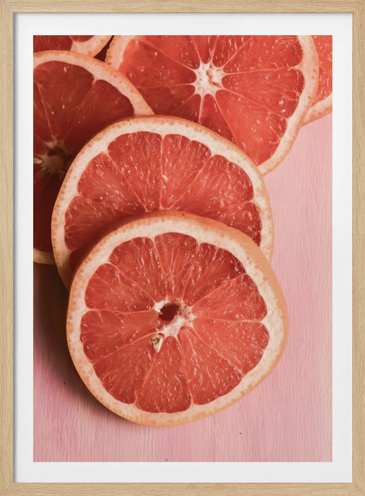 A close-up photograph of several overlapping slices of vibrant pink grapefruit arranged on a pale pink surface, enclosed within a simple black frame. Print