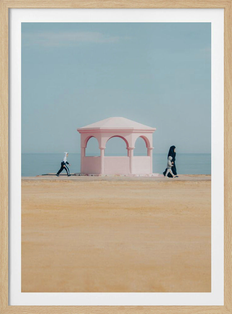 A minimalist photograph of a coastal scene with a pastel color palette. A pink arched gazebo sits on a promenade with the blue sea and sky in the background. A large expanse of beige sand is in the foreground. Two figures in hijabs walk in opposite directions on either side of the gazebo, one with a small child. Decor