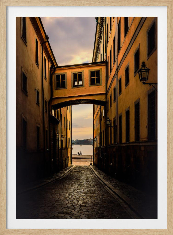 A perspective view down a narrow, shadowy cobblestone alley in an old European city, framed by warm, ochre-colored buildings. An arched walkway connects the buildings overhead, and the alley opens to a view of a tranquil body of water at sunset, with two small figures silhouetted by the shore. Decor