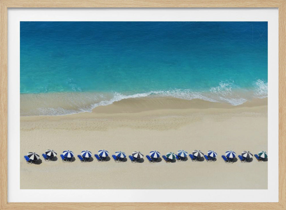 An aerial view of a pristine sandy beach with vibrant turquoise ocean waves gently lapping the shore. A long, neat row of blue and white striped beach umbrellas stands along the sand, creating a serene and orderly coastal scene, all enclosed within a silver frame. Artwork
