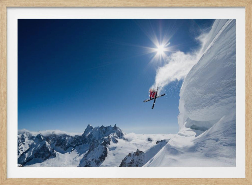 An extreme skier in a red jacket is captured mid-air, having just launched off a steep, snowy cornice, kicking up a cloud of powder against a brilliant blue sky with a bright sunburst overhead and a panoramic view of snow-capped mountains below. Poster