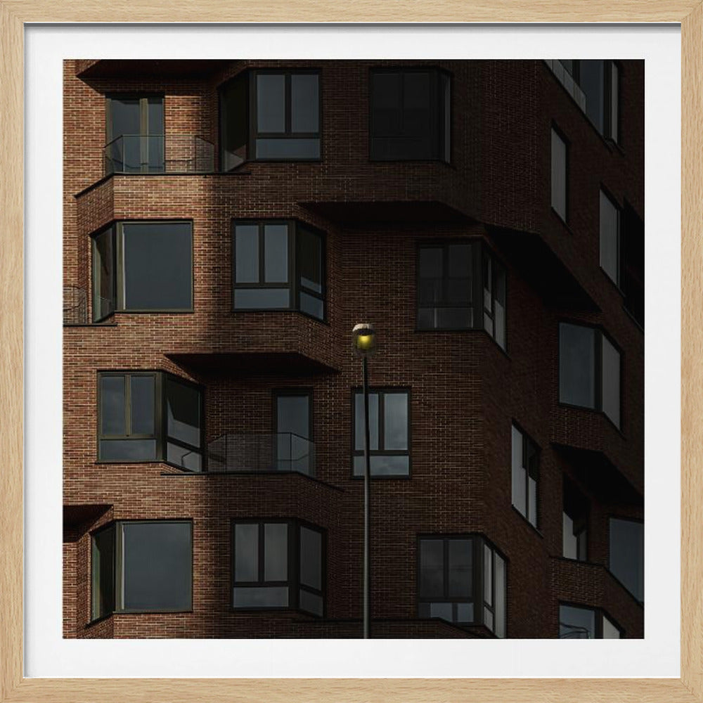 A close-up photograph of a modern red-brick apartment building, framed in light wood. A sharp shadow line divides the facade, with a lit streetlamp providing a warm yellow glow in the shaded area. Decor