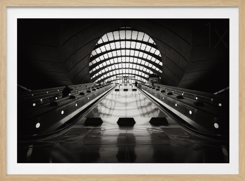 A dramatic low-angle, black and white photograph of the escalators at Canary Wharf station. The perspective looks up the sleek, reflective escalators towards a massive, arched, grid-patterned window at the far end, creating strong leading lines and a sense of scale within the modern, curved architecture. Decor