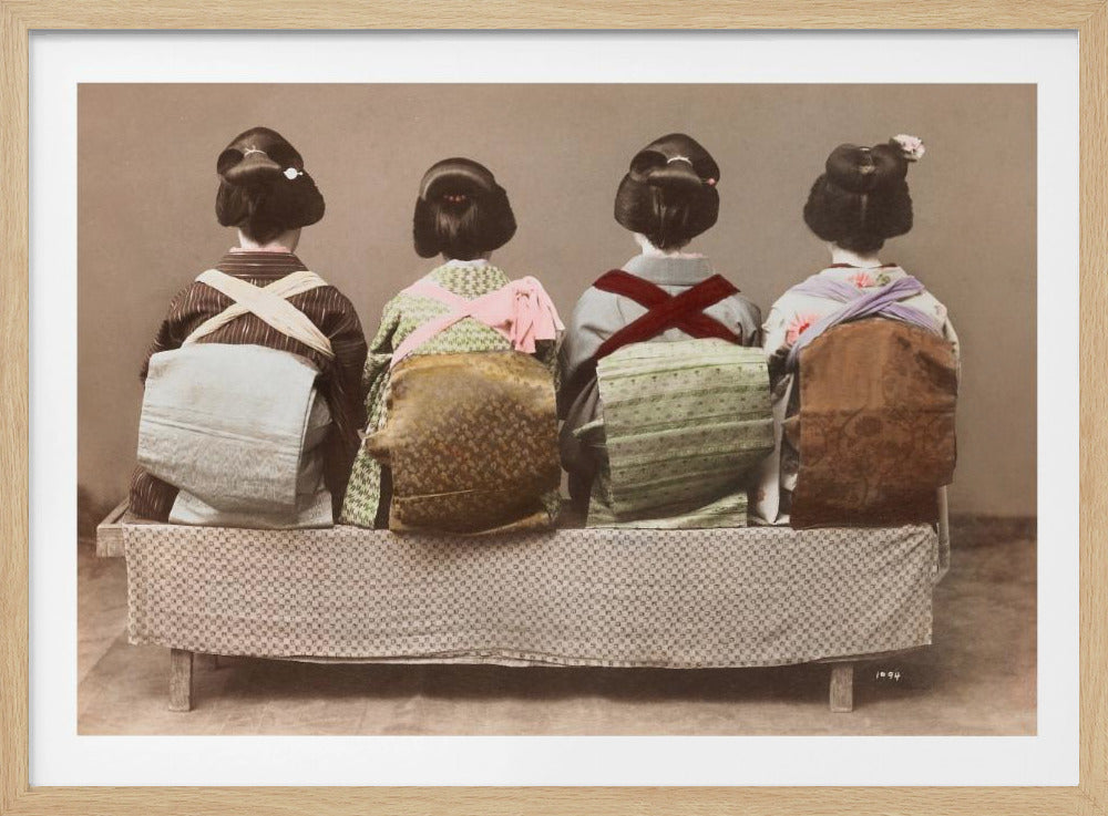 A framed, vintage, hand-colored photograph showing the backs of four Japanese women wearing traditional kimonos and with elaborate updos, sitting in a row on a patterned bench against a plain sepia background. Print