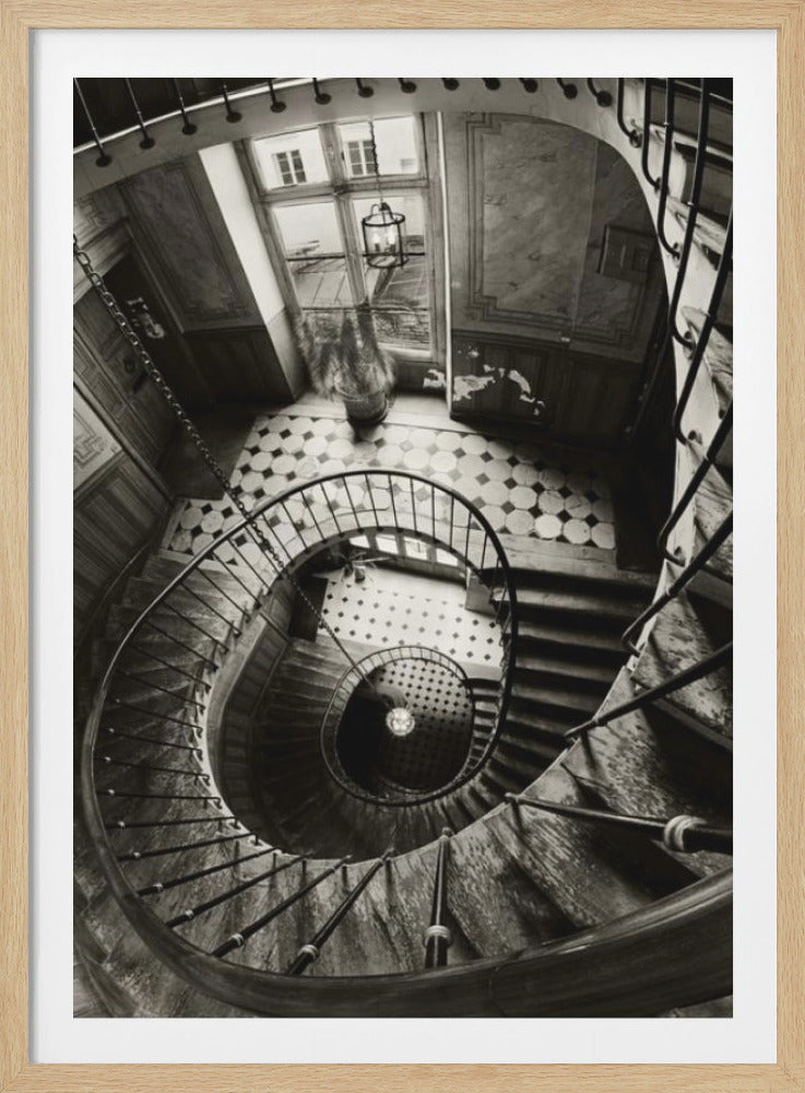 A dramatic, high-angle black and white photograph looking down a vintage spiral staircase. The elegant metal railing spirals downwards, creating a vortex-like perspective. At the top, a landing with a checkered floor is lit by a large window. Wall Art