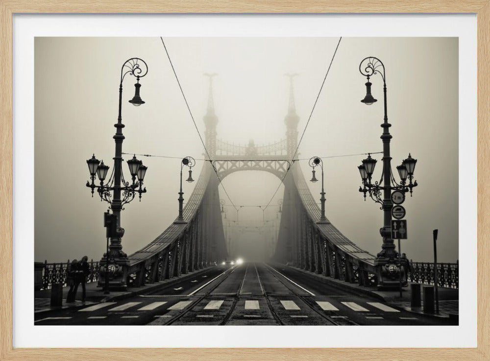 A framed, symmetrical black and white photograph of a large, ornate metal bridge disappearing into thick fog. Vintage lampposts stand on either side in the foreground, and the road and tram tracks lead the eye into the misty distance where faint car headlights glow. Print