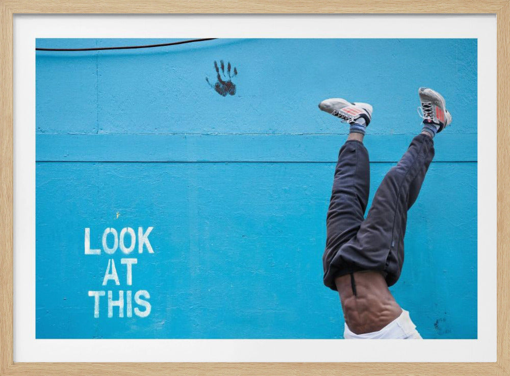A person's lower body doing a handstand against a bright blue wall with the stenciled words 'LOOK AT THIS' in white. The person wears dark pants and sneakers, and their muscular abdomen is visible. A black handprint is on the wall above the text. The artwork is presented in a silver frame. Print