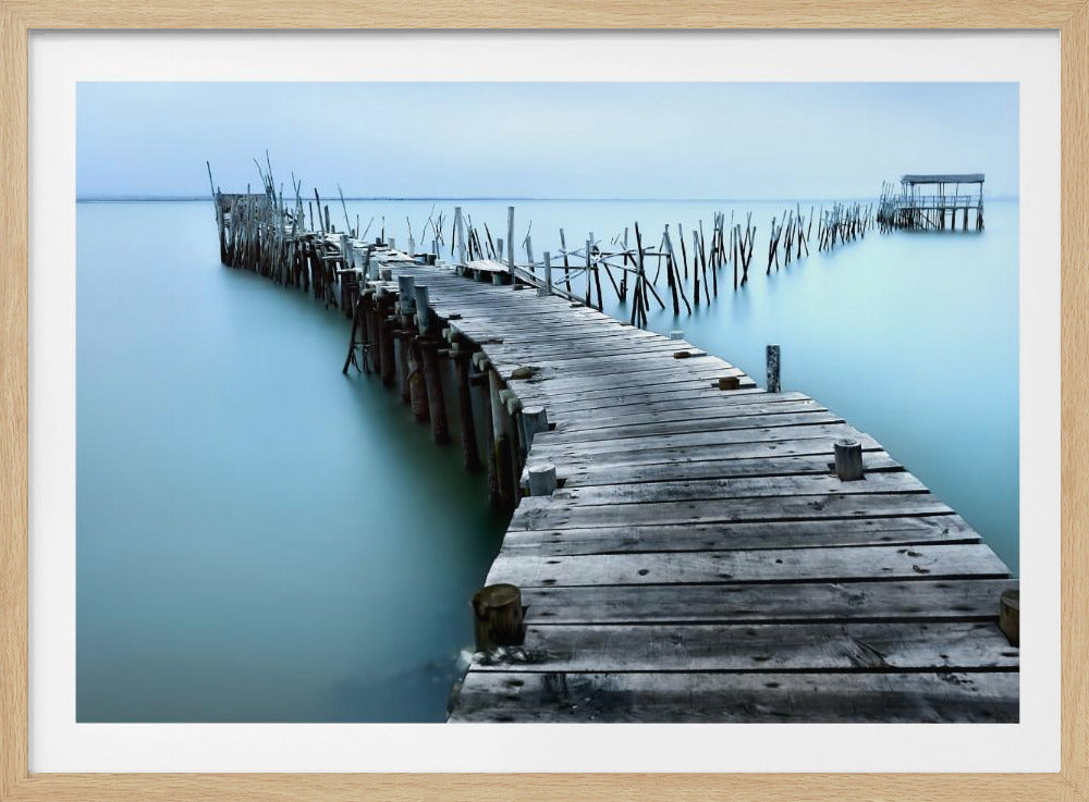 A serene long-exposure photograph of a weathered wooden pier curving over calm, misty blue water towards a distant fishing hut under a pale blue sky, all enclosed in a silver frame. Poster