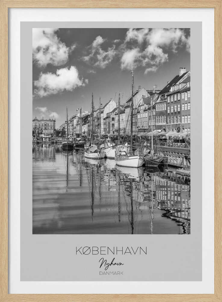 A black and white poster of Nyhavn harbor in Copenhagen. The photograph shows traditional sailboats and ships moored along a canal, with a line of historic, multi-story buildings on the far bank. The sky has fluffy clouds, and the entire scene is reflected in the calm water. The poster has a white border with the text 'KØBENHAVN', 'Nyhavn', and 'DANMARK' at the bottom, and is shown within a black frame. Poster