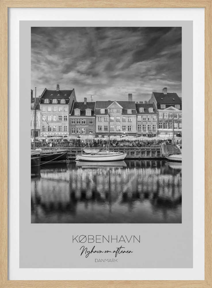 A framed black and white photograph of the Nyhavn harbor in Copenhagen, Denmark. The image shows a row of classic gabled houses along the waterfront, with several sailboats moored in the canal. The buildings and a dramatic cloudy sky are reflected in the water. At the bottom, a white border contains the text 'KØBENHAVN', 'Nyhavn om aftenen', and 'DANMARK'. Wall Art