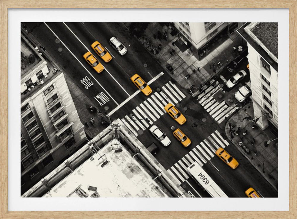 An aerial, black and white photograph of a bustling New York City intersection, with the iconic yellow taxi cabs highlighted in a vibrant selective color. Pedestrians cross the street as cars and a bus navigate the busy roads between tall buildings. Wall Art