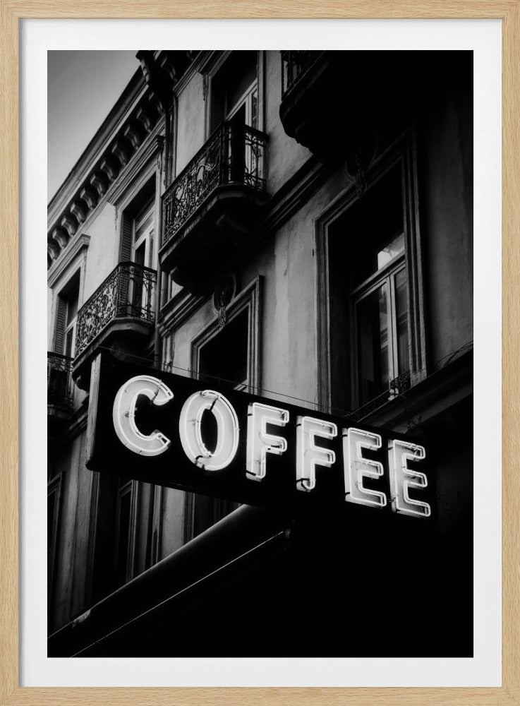 A dramatic, low-angle, black and white photograph of a bright white neon sign that reads 'COFFEE', mounted on a classic-style building with ornate balconies. Print